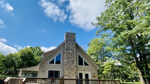 Une maison à deux étages avec une cheminée en pierre, une terrasse en bois et un grand arbre en arrière-plan.