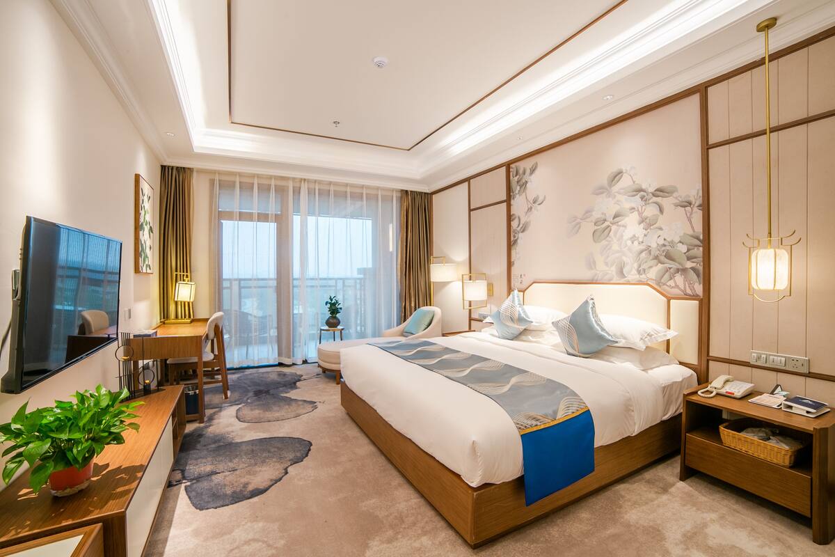 A hotel room with a large bed, a desk with a chair, a TV, and a potted plant.