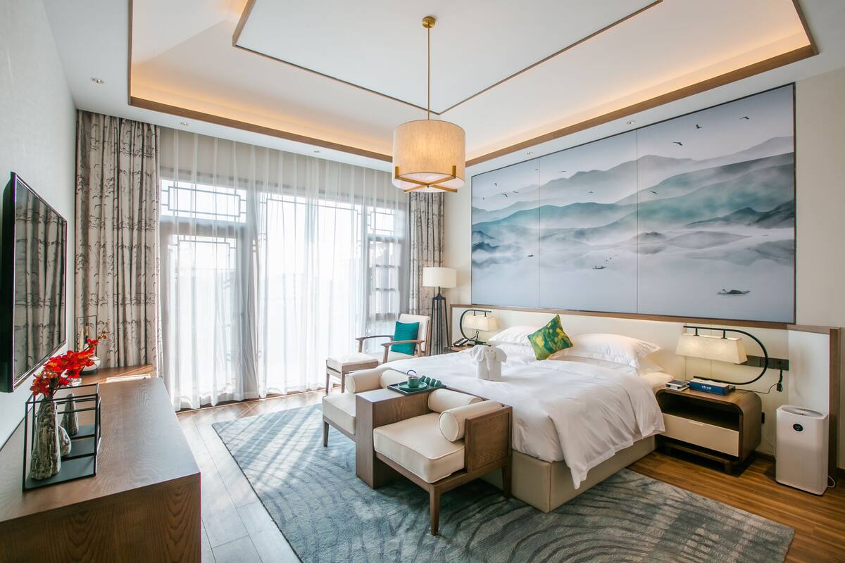 A modern hotel room with a large bed, a desk, a chair, a nightstand, and a large painting on the wall.