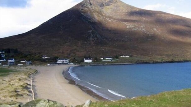 Eine Küstenlandschaft mit Sandstrand, einer kleinen Siedlung mit Häusern und einem markanten Hügel oder Berg im Hintergrund.
