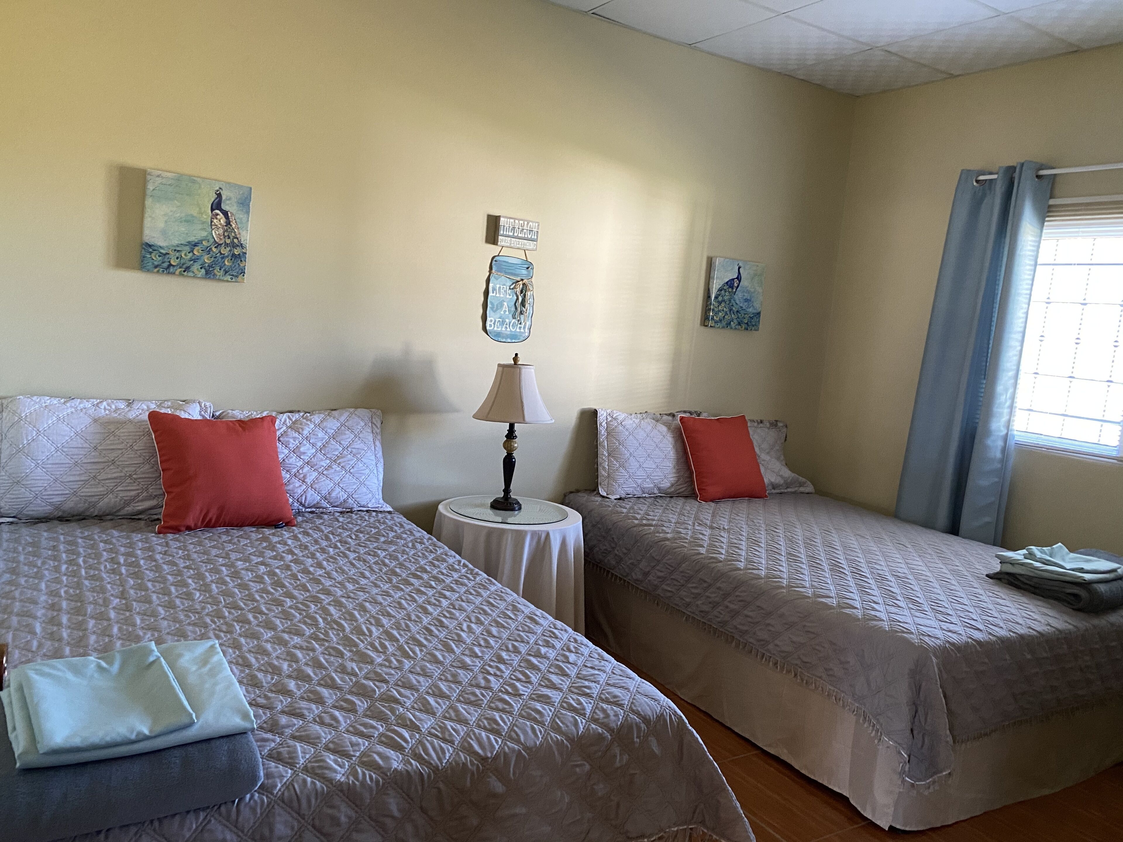 Habitación de hotel con dos camas, cada una con colcha acolchada y almohadas, una mesita redonda con lámpara y obras de arte en las paredes.