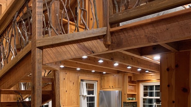A wooden cabin interior with a kitchen, dining area, and a staircase leading to a loft.