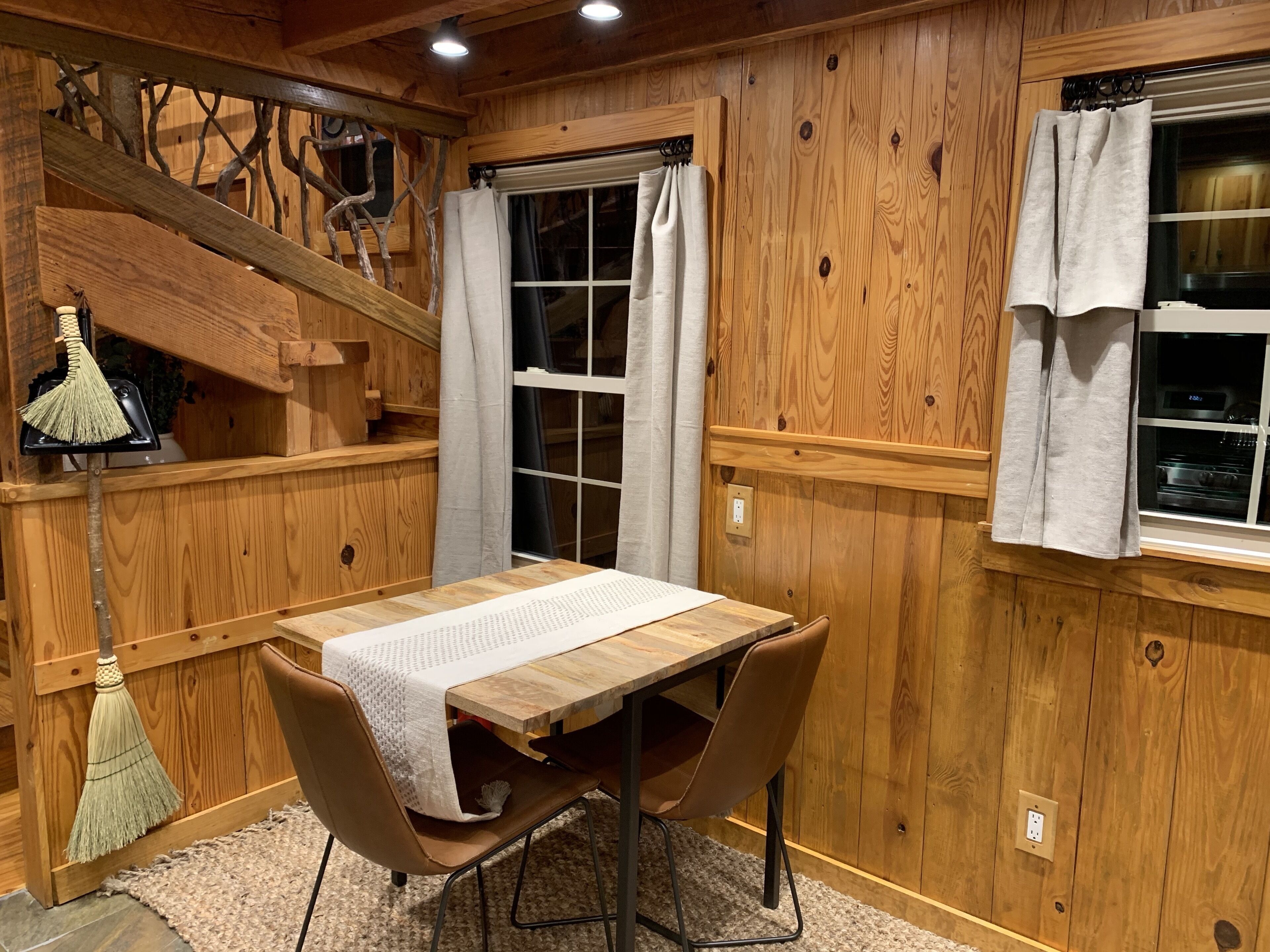 A wooden-paneled room with a dining table, chairs, a broom, and a window with curtains.