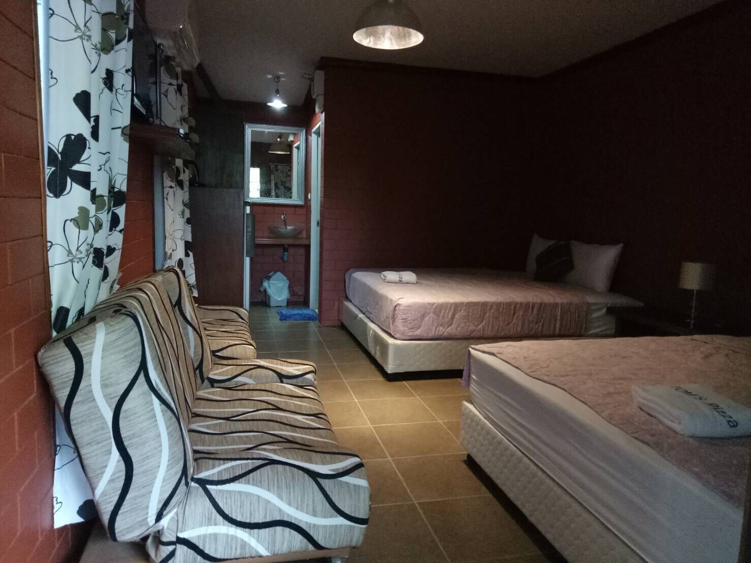 A hotel room with a sofa, two beds, a bathroom with a sink, and a hallway leading to another room.