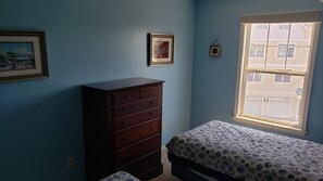 Une chambre à coucher comprenant un lit, une commode, deux tableaux encadrés et une fenêtre donnant sur des immeubles.