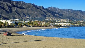 Una playa con montañas al fondo, cielos despejados y una hilera de edificios blancos a lo largo de la costa.