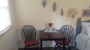 Un pequeño comedor con una mesa y dos sillas, una ventana con persianas, y una lámpara en la pared.