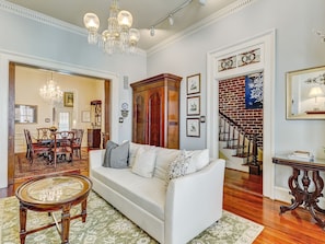 A spacious living room with a white sofa, a round coffee table, and a chandelier. There is a dining area with a wooden table and chairs, and a staircase with a brick wall.