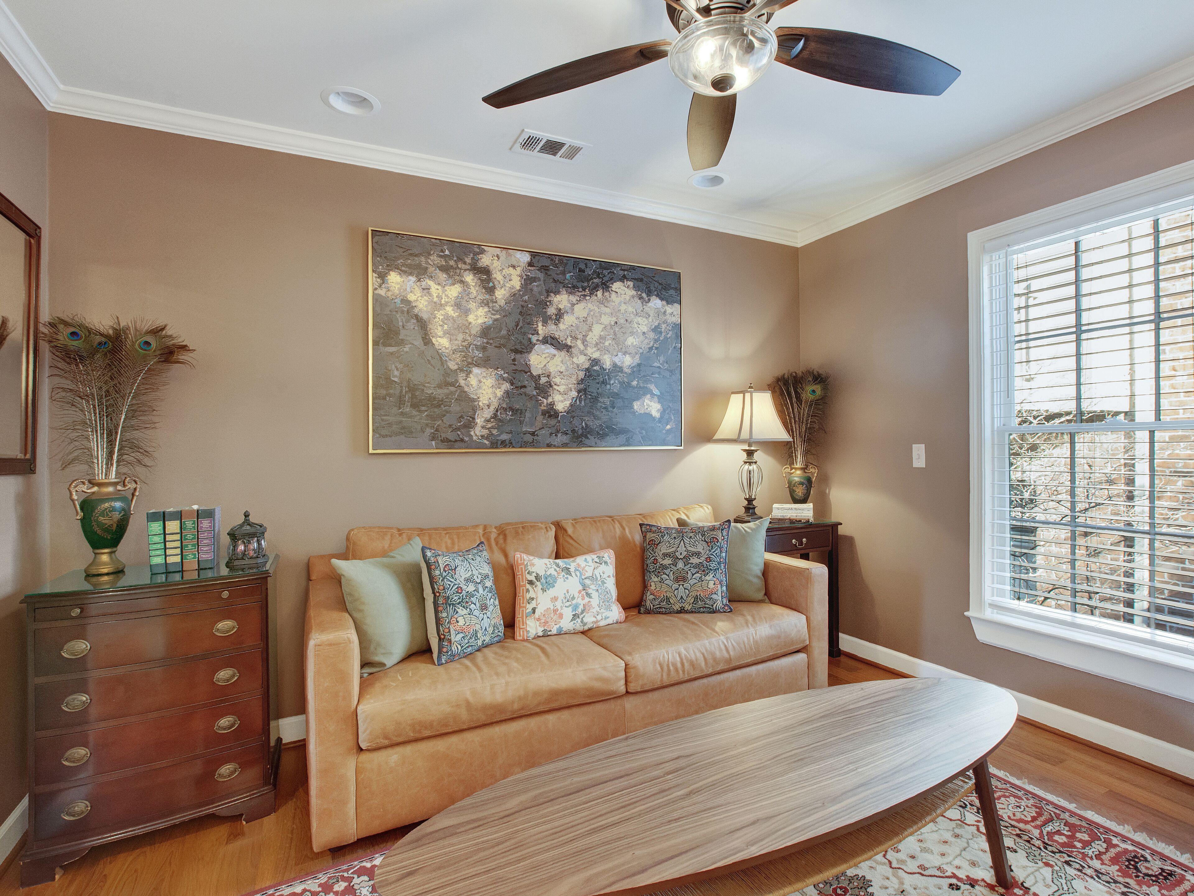 A living room with a beige sofa, decorative pillows, a wooden coffee table, a world map painting, a floor lamp, and a window with blinds.