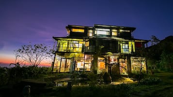 A multi-story house with large windows and a balcony, surrounded by vegetation.