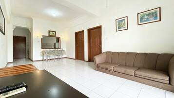 A spacious living room with a brown sofa, a dining table with chairs, and a black coffee table. There are two wooden doors and a framed picture on the wall.