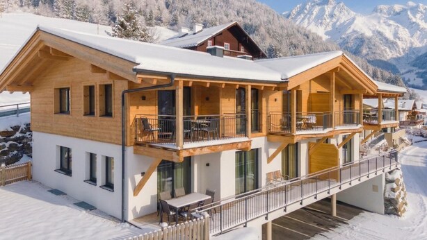 A wooden chalet with a balcony, large windows, and a view of snow-covered mountains.
