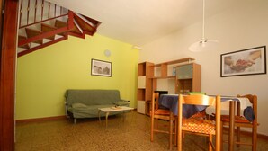 A cozy living room with a green sofa, wooden dining table, and chairs. A staircase with wooden railings is visible in the background. - Delta Blu Residence Village - Villetta Trilocale 05 (Lido di Pomposa)