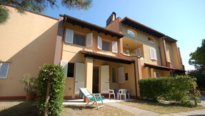 A two-story building with a balcony, a small garden area with a chair and table, and a potted plant. - Delta Blu Residence Village - Villetta Trilocale 05 (Lido di Pomposa)