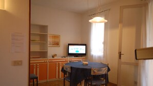 A small dining area with a table and chairs, a television on a stand, and a window with curtains. - Delta Blu Residence Village - Villetta Trilocale 16 (Lido di Pomposa)