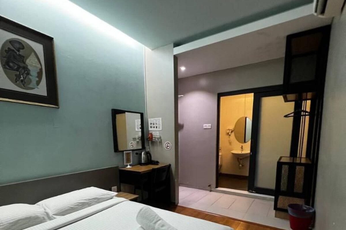 Une chambre d’hôtel avec un lit, un bureau, une chaise, un miroir et une salle de bain visible à travers une porte ouverte.