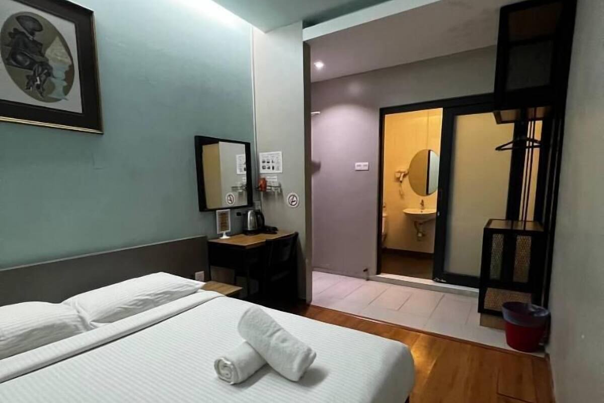 Une chambre d’hôtel avec un lit, un bureau, une chaise, un miroir et une salle de bain visible à travers une porte ouverte.