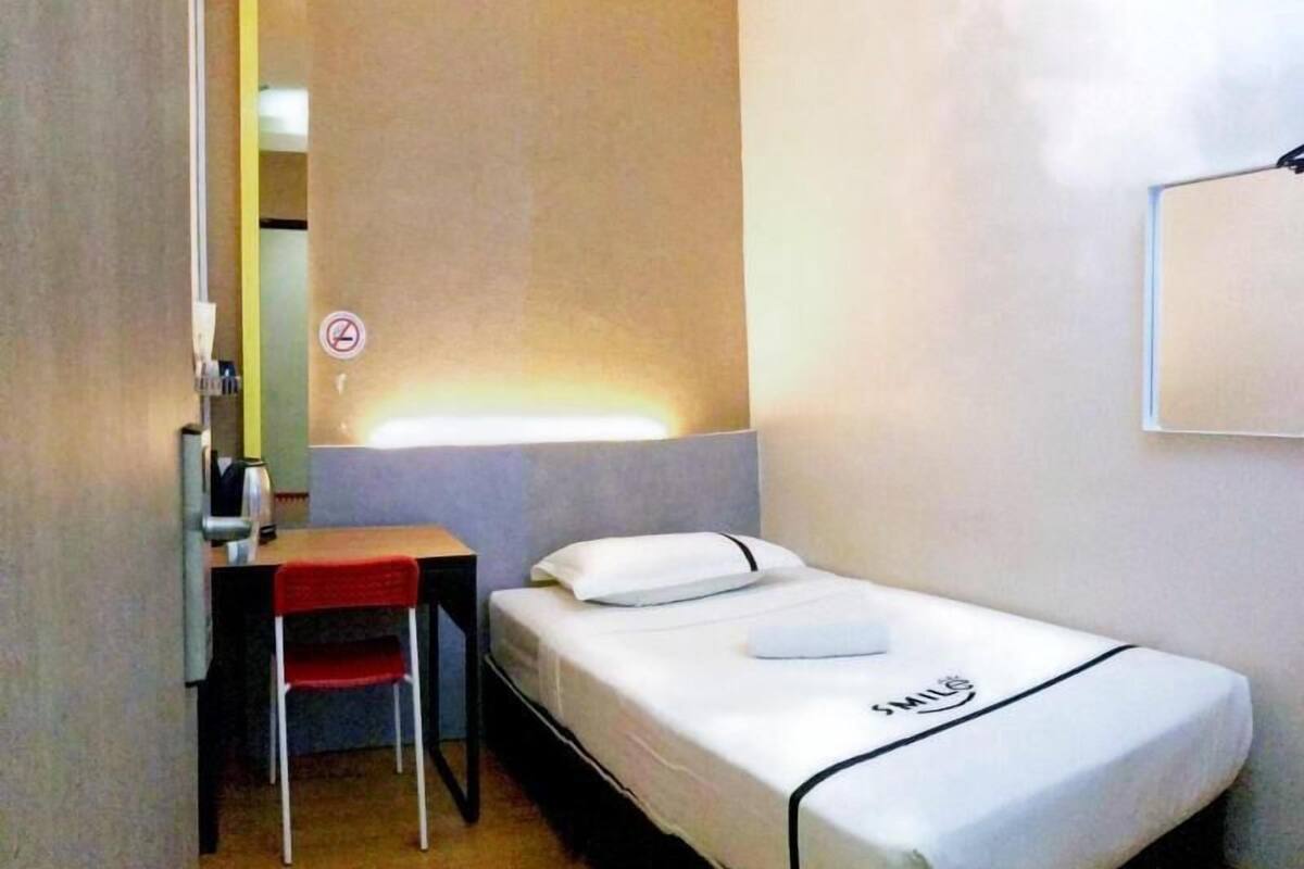 Une petite chambre avec un lit simple, un bureau et une chaise, une table de chevet et un miroir fixé au mur.