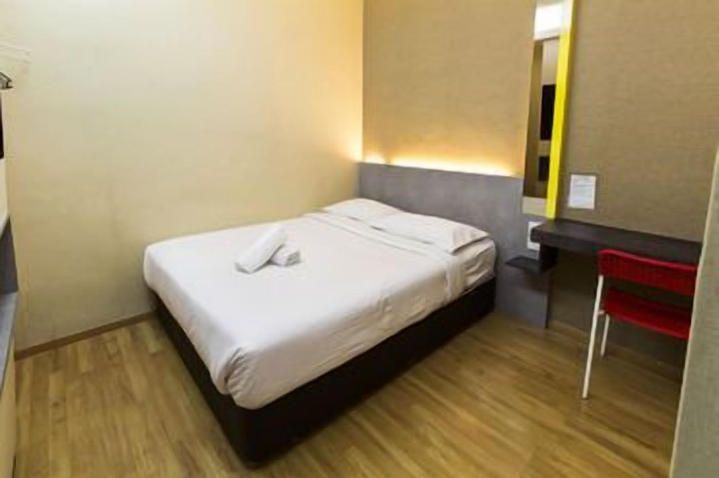 A small, neatly made bed with a white duvet and a single pillow, a wooden floor, a small bedside table with a red chair, and a mirror on the wall.