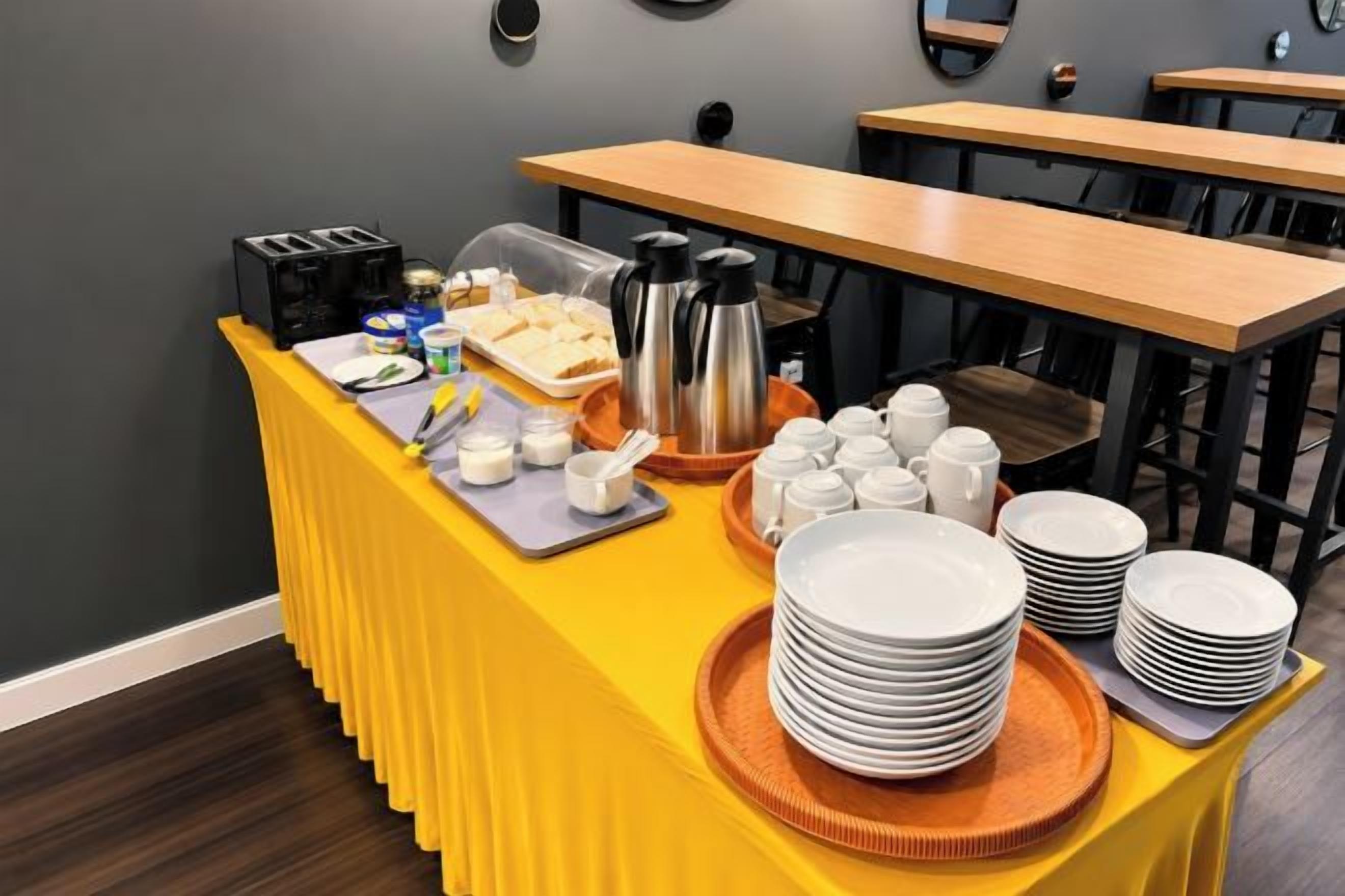 A buffet setup with a yellow table, plates, cups, and a coffee pot.