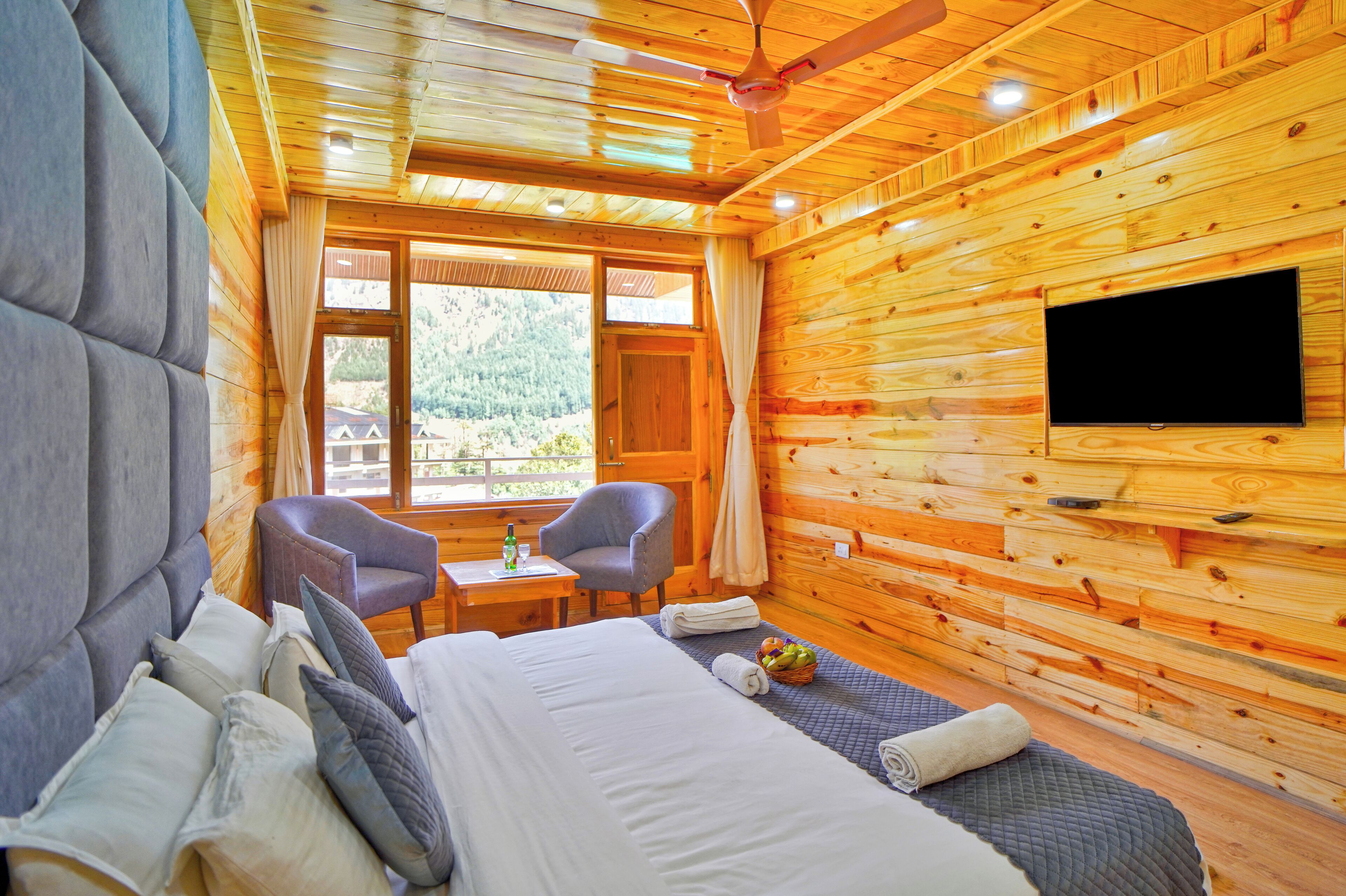 A wooden cabin room with a bed, two chairs, a small table, and a TV on the wall.