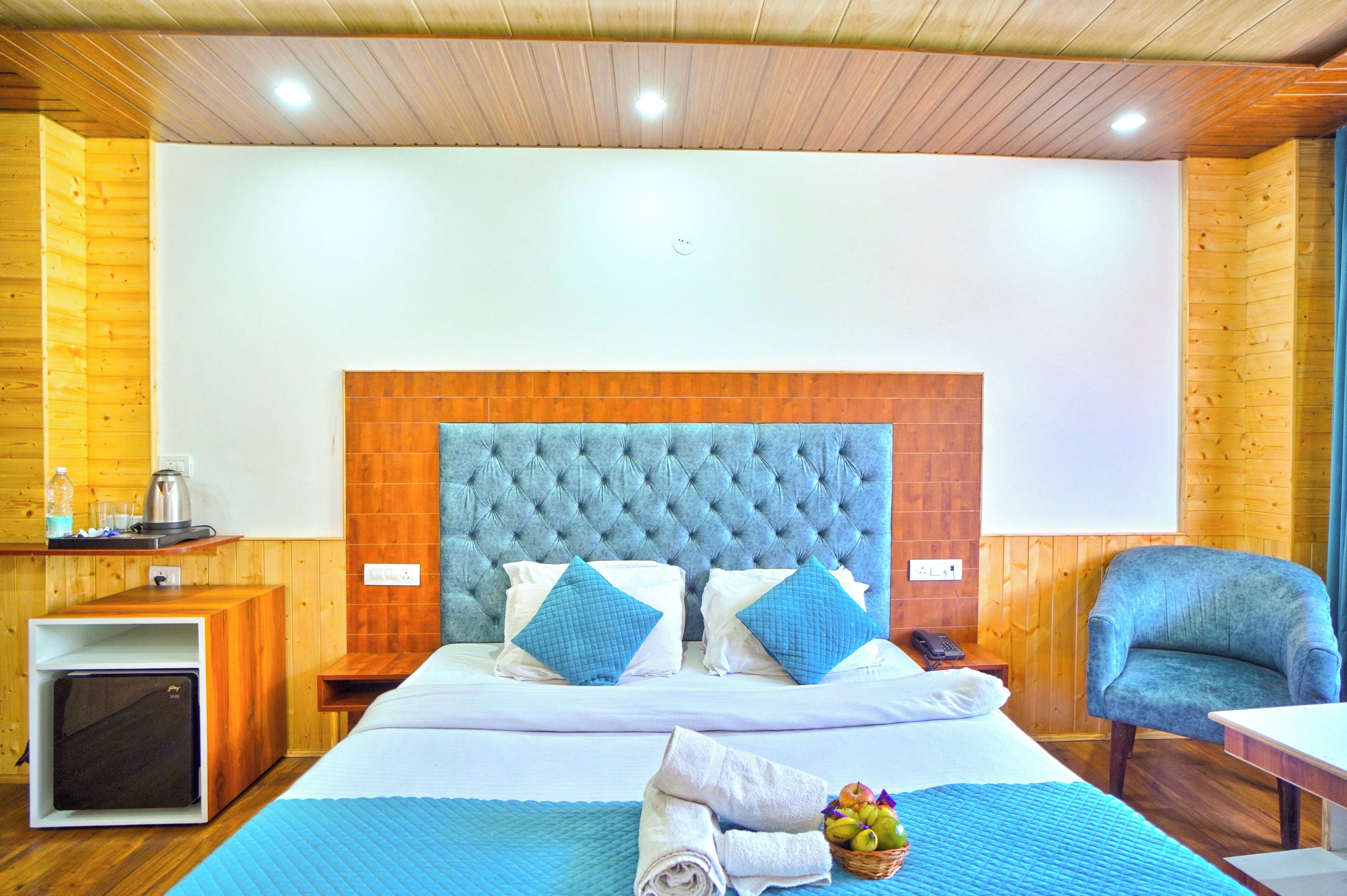 A hotel room with a large bed, wooden walls, a blue upholstered headboard, and a small desk with a lamp.