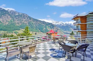 Una terraza con mobiliario de mimbre y piso a cuadros, con vistas a un paisaje montañoso de picos nevados.
