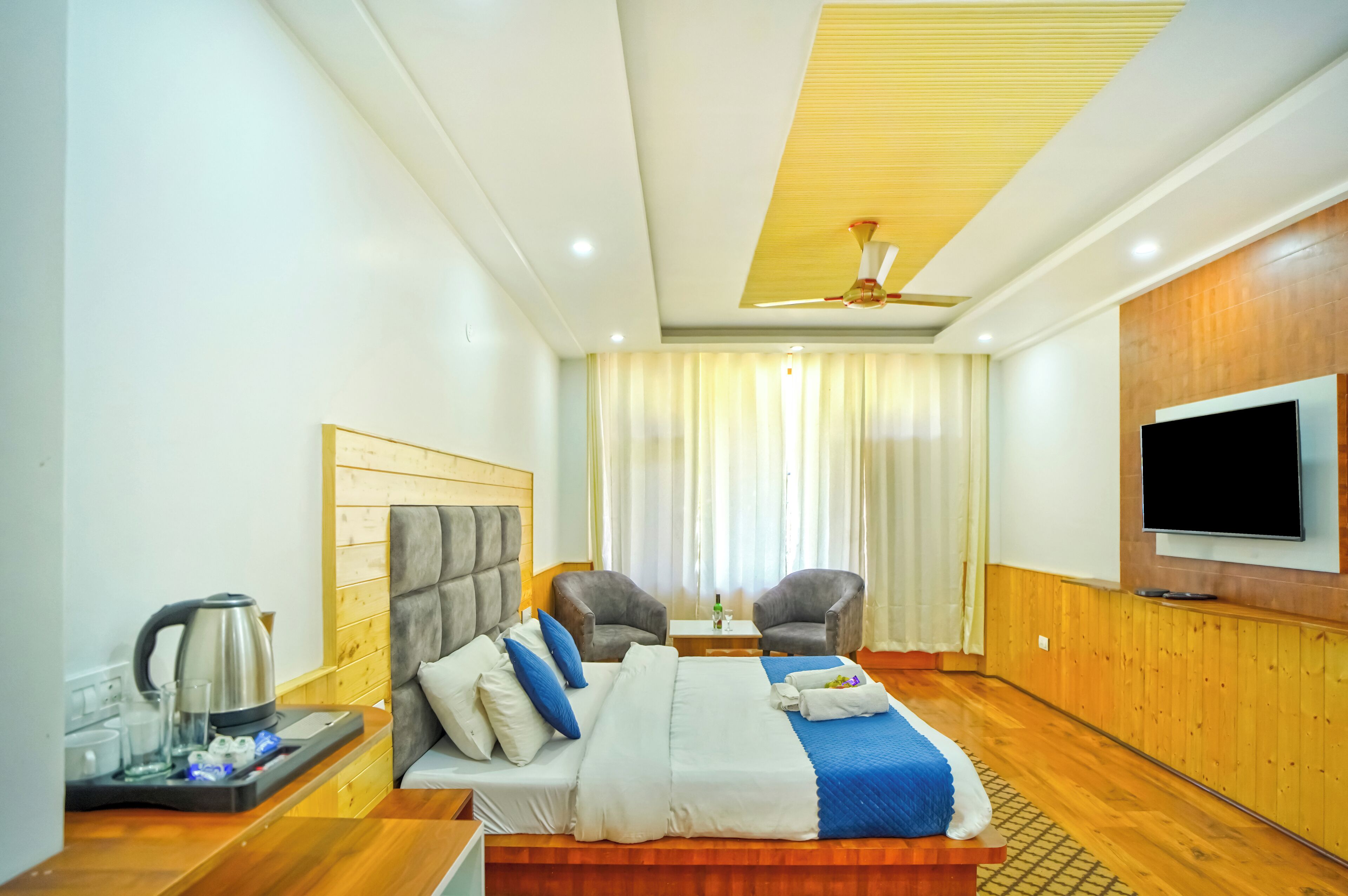 A hotel room with a bed, two armchairs, a TV, a ceiling fan, and a wooden floor.