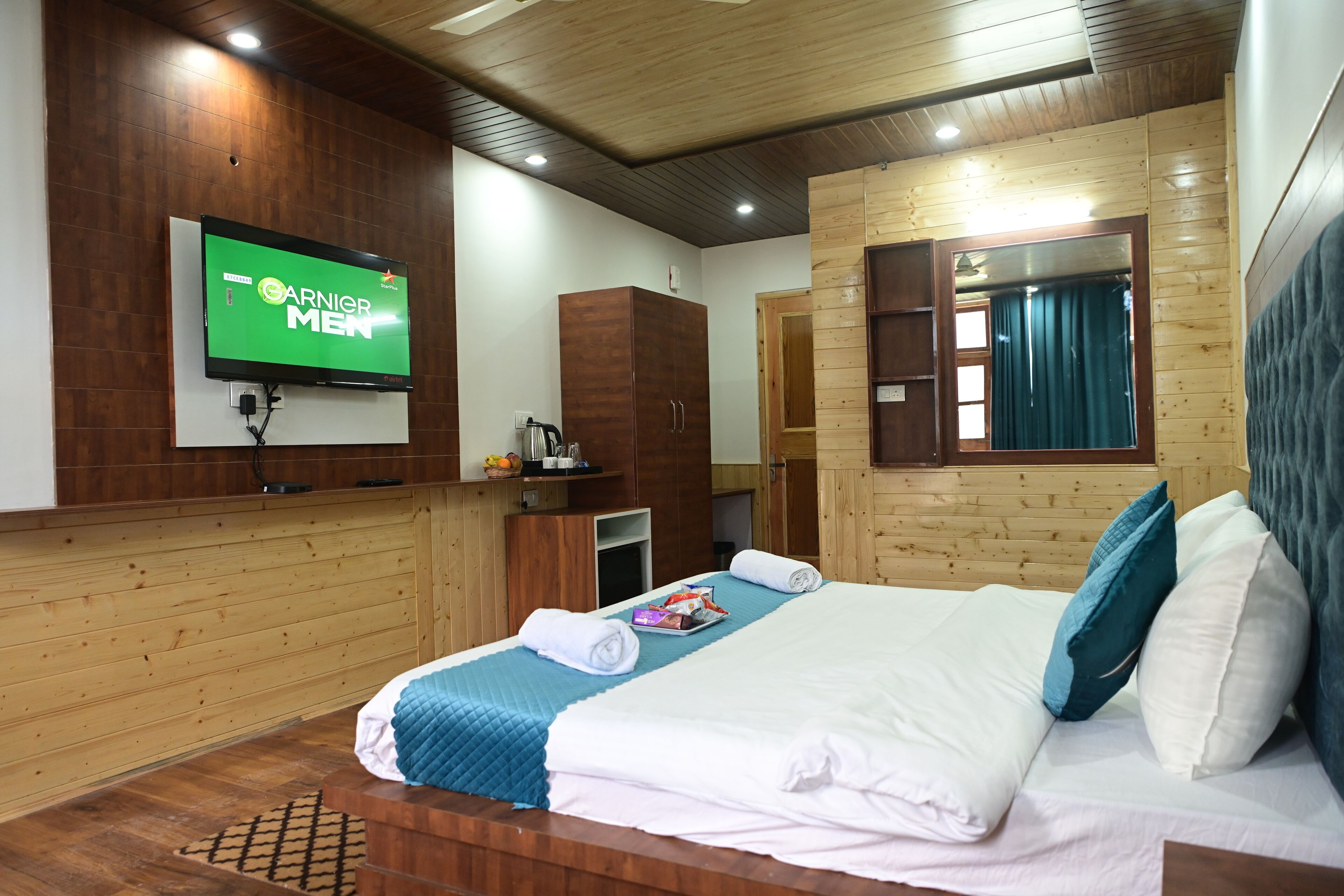 A hotel room with a wooden interior, a television mounted on the wall, a bed with white and blue bedding, and a wardrobe.