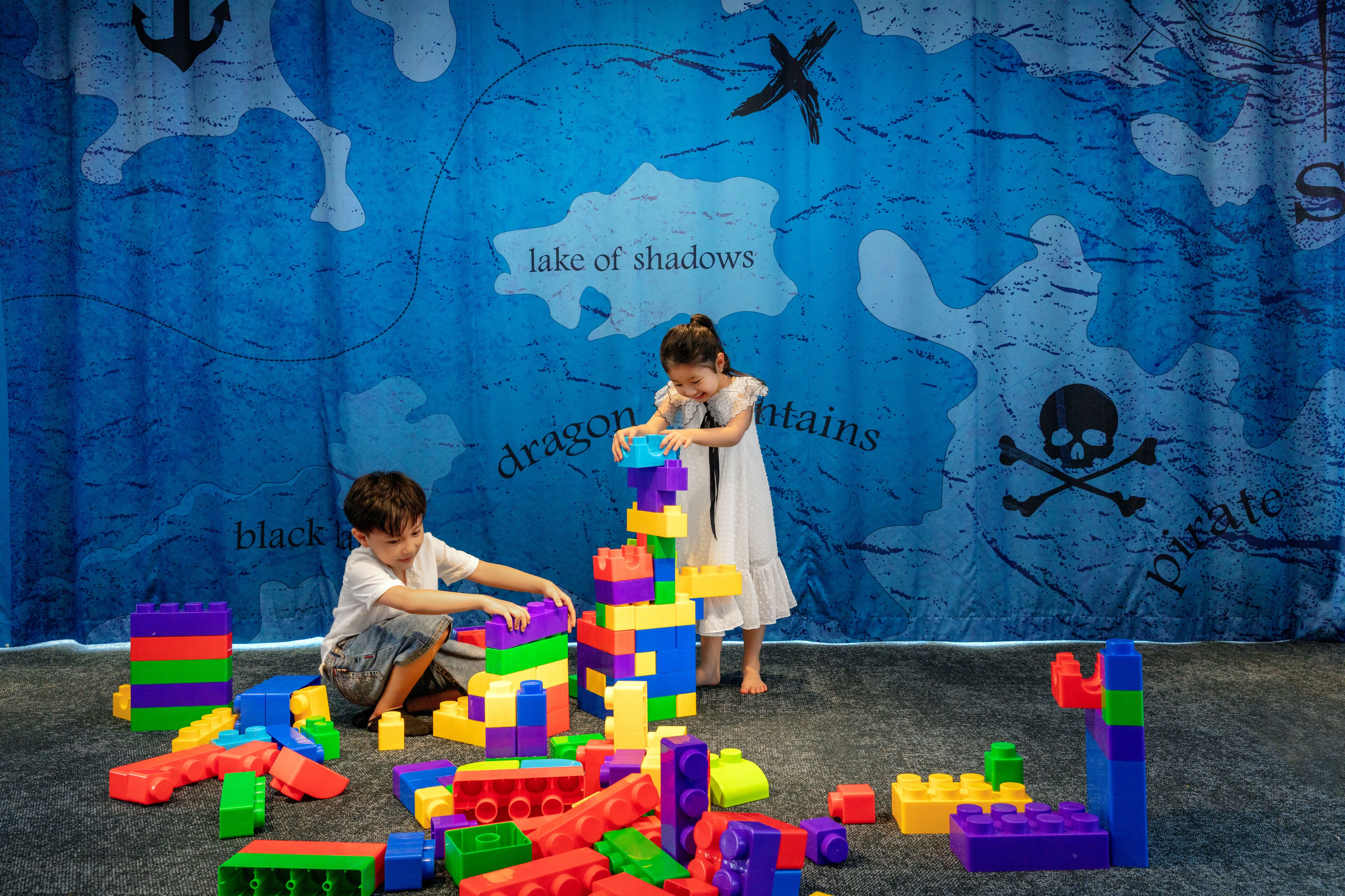 Children playing with colorful building blocks in front of a blue backdrop with maritime-themed designs.