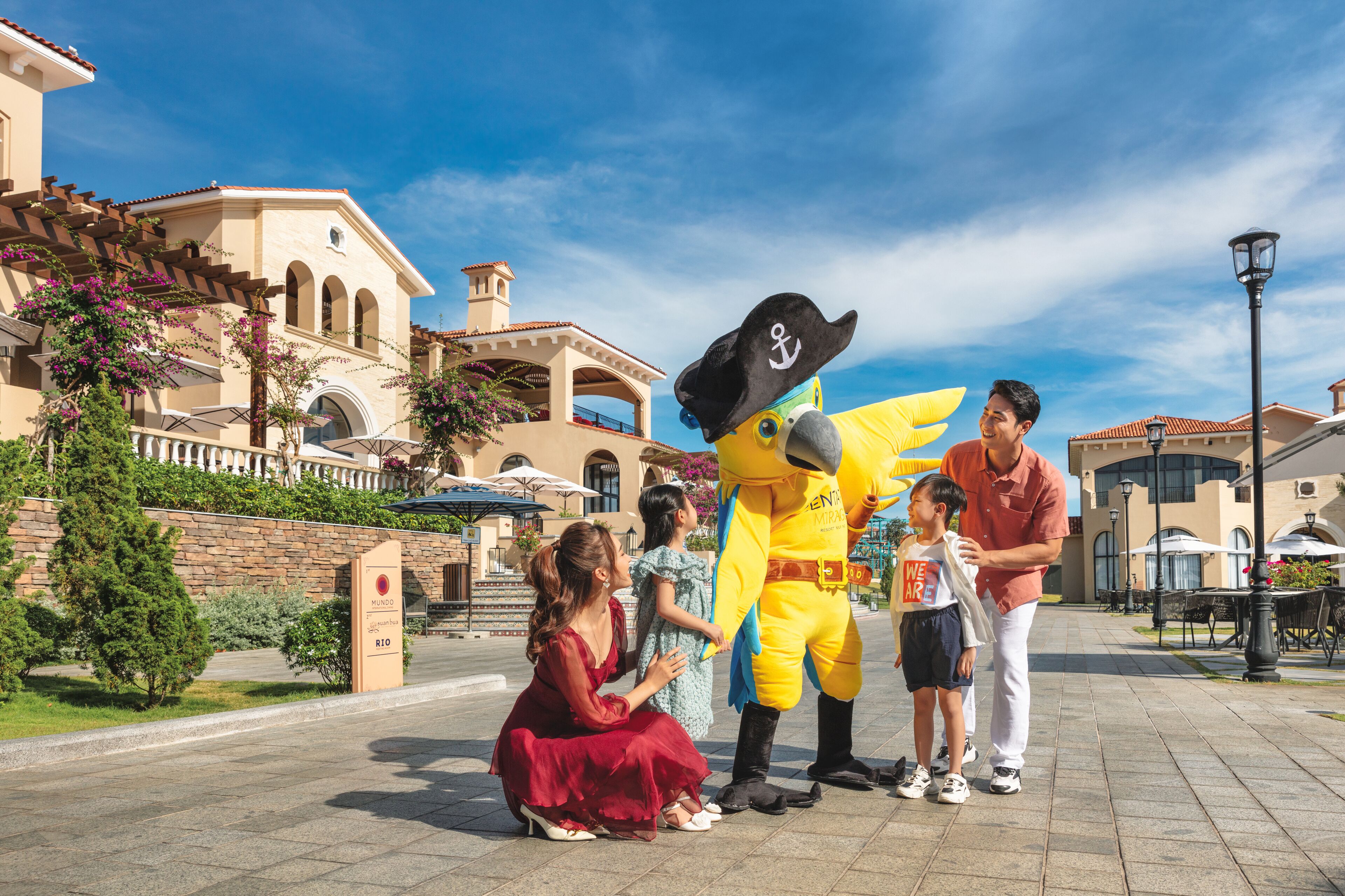 Une mascotte interagissant avec des visiteurs sur une place dotée d’une architecture de style méditerranéen, sous un ciel dégagé.