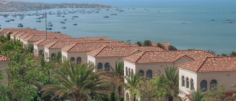 A coastal hotel with terracotta roofs, palm trees, and a view of the sea with numerous boats.