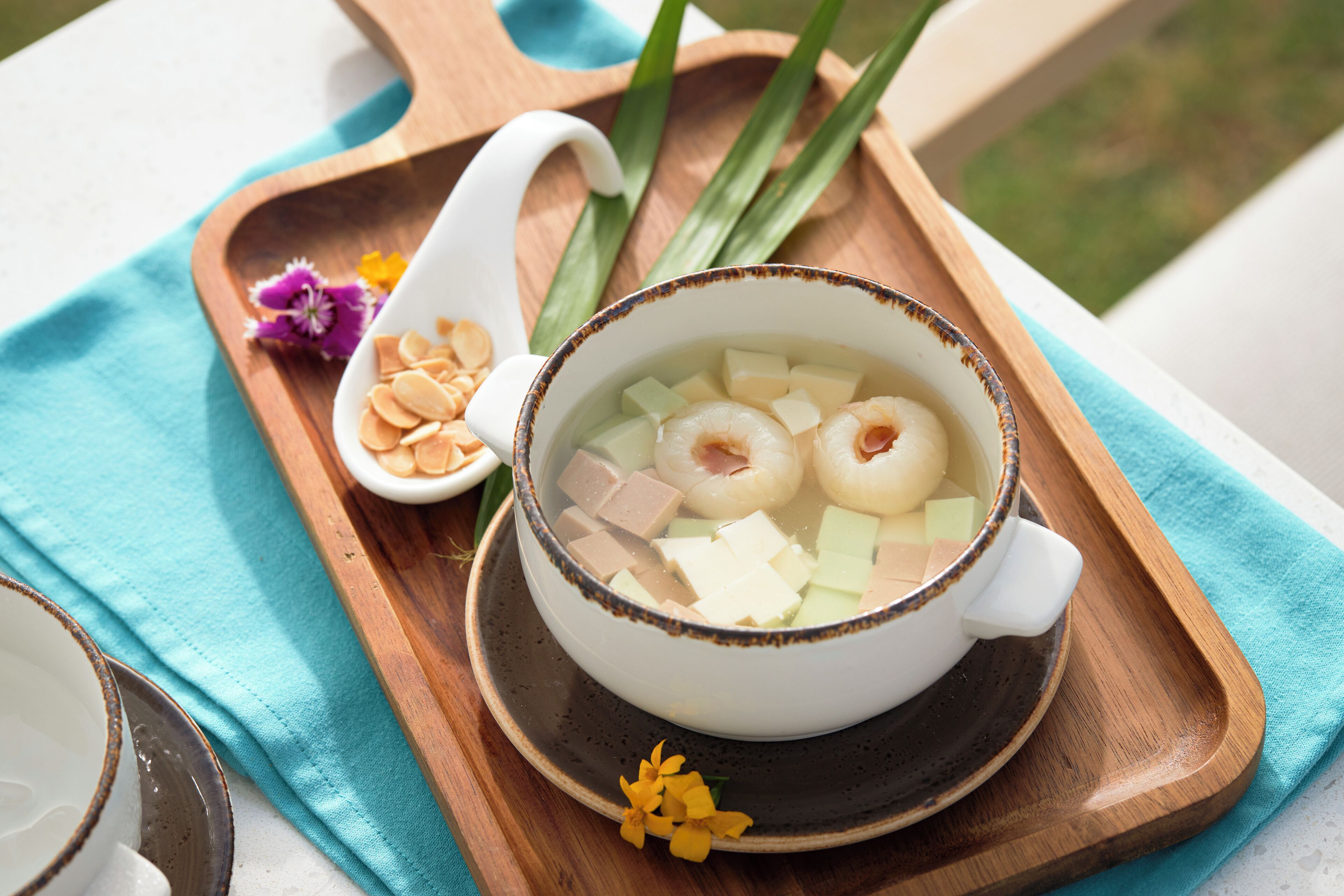 Un tazón de sopa con tofu y vieiras sobre una bandeja de madera con un paño azul.
