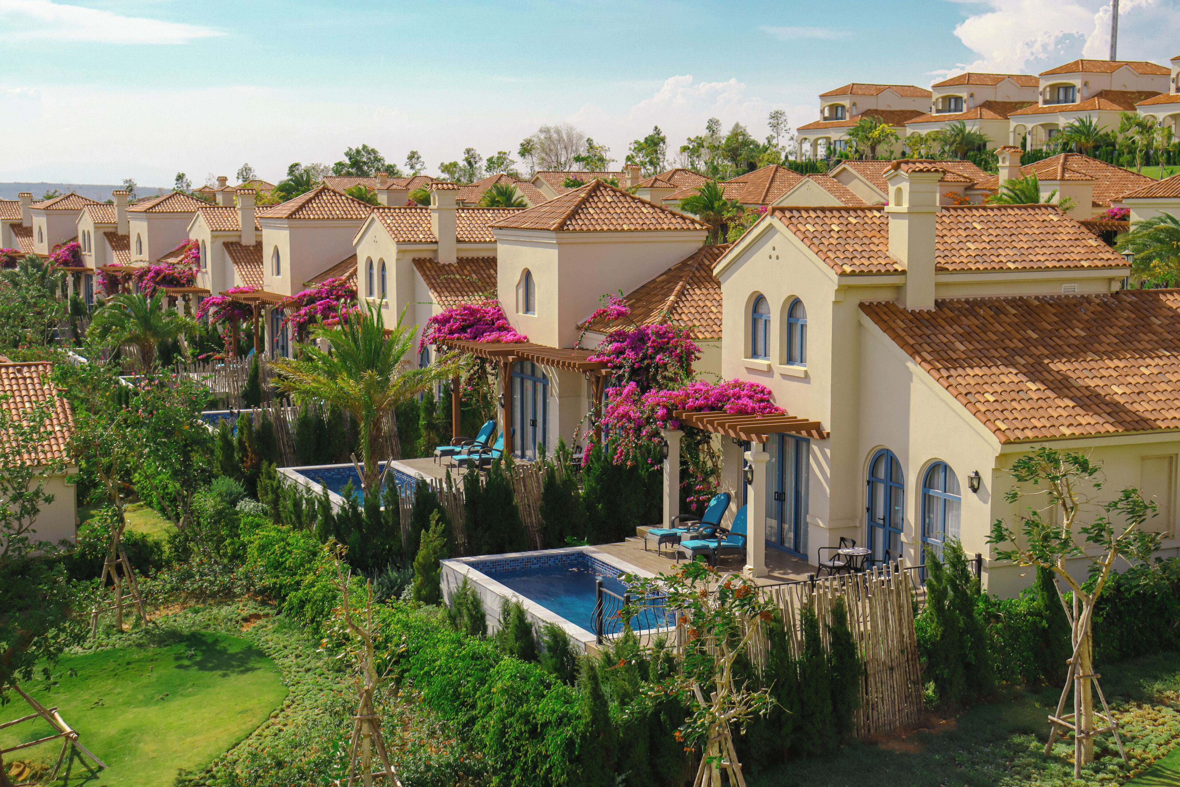 A row of Mediterranean-style villas with swimming pools, lush gardens, and a clear blue sky.
