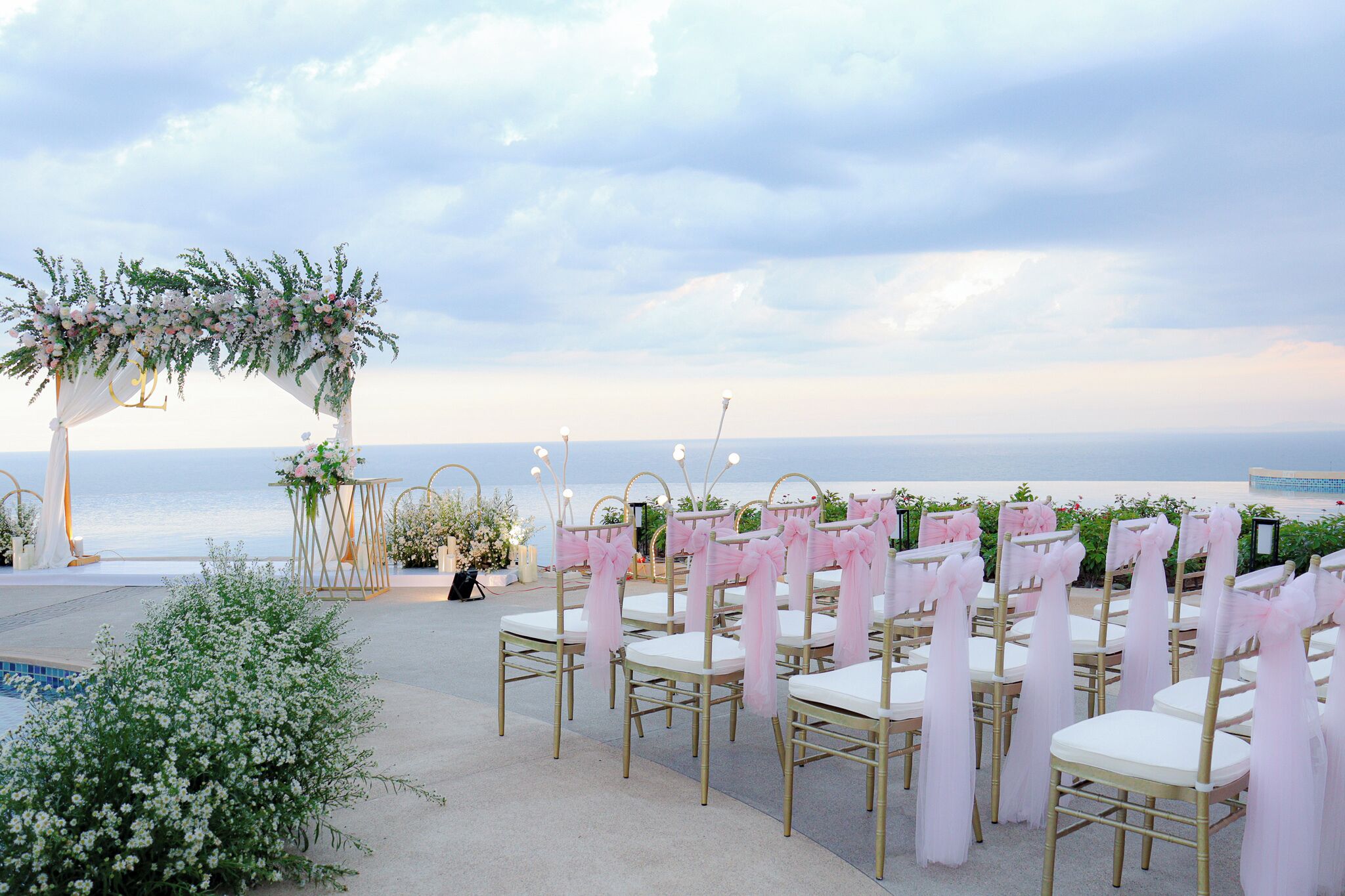Hochzeitszeremonie im Freien mit Blick auf den Ozean, mit Reihen von Stühlen mit weißen Kissen und goldenen Rahmen, verziert mit rosa Bändern, und einem Blumengestell.