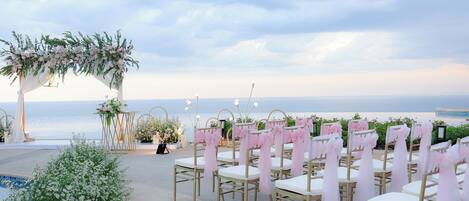 An outdoor wedding setup with a view of the ocean, featuring rows of chairs with white cushions and gold frames, adorned with pink sashes, and a floral archway.