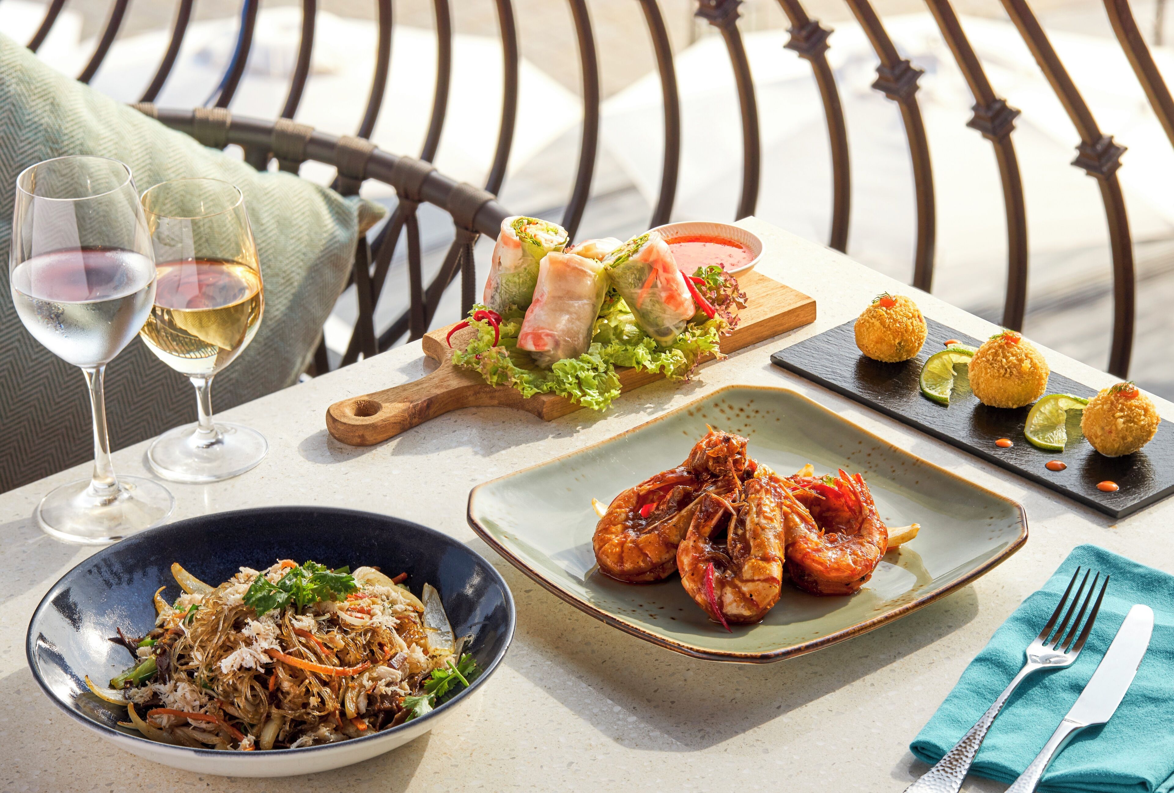 A table set with various dishes, including shrimp, spring rolls, and a salad, along with two glasses of white wine.