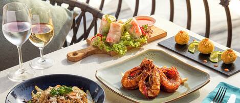 A table set with various dishes, including shrimp, spring rolls, and a salad, along with two glasses of white wine.