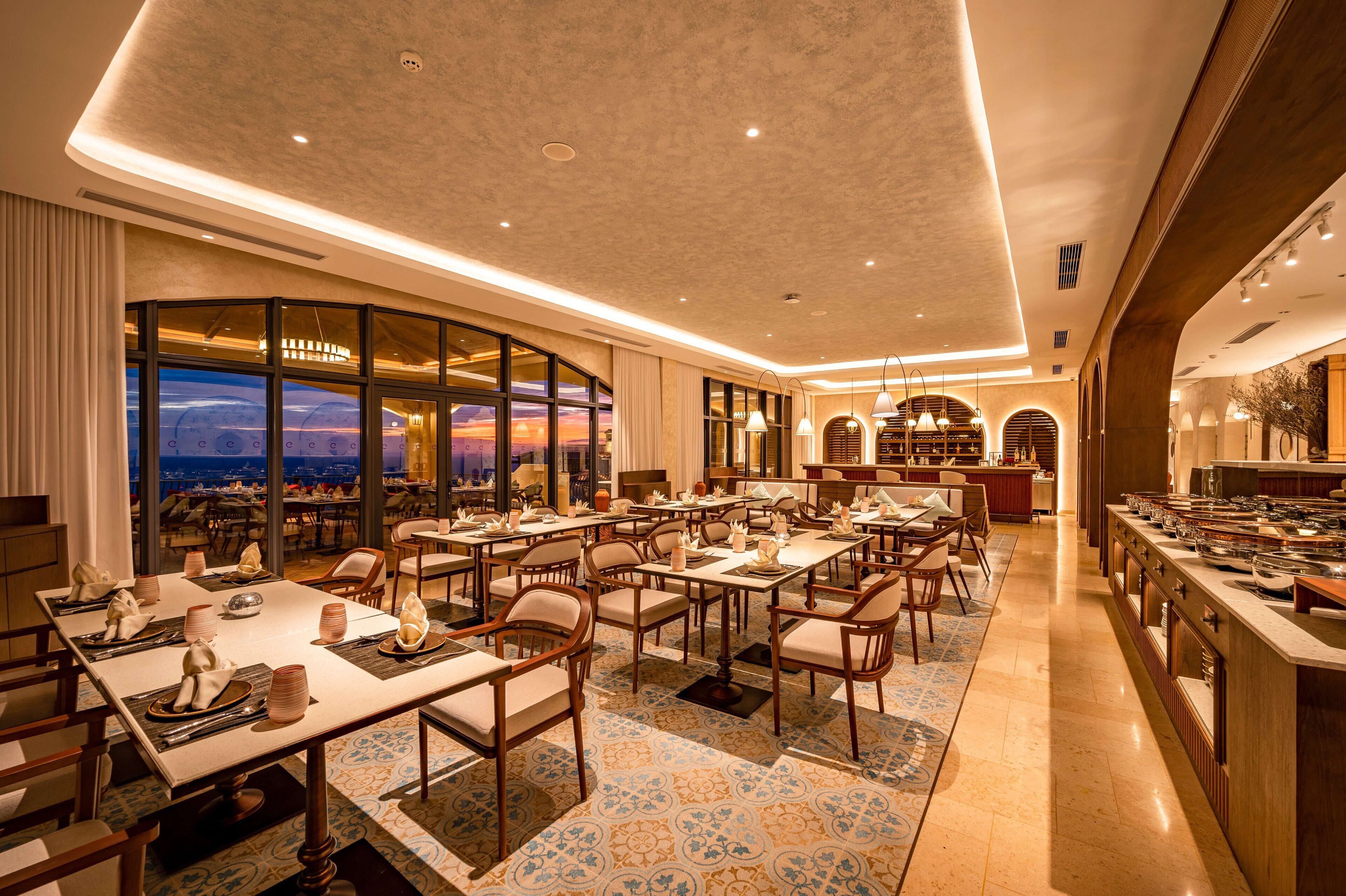 Un vaste espace dédié à la salle à manger, avec plusieurs tables et chaises, une grande fenêtre offrant une vue sur le coucher du soleil, et une cuisine moderne visible au fond.