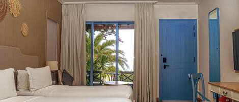 A hotel room with two beds, a balcony with curtains, a blue door, and a desk with a chair.