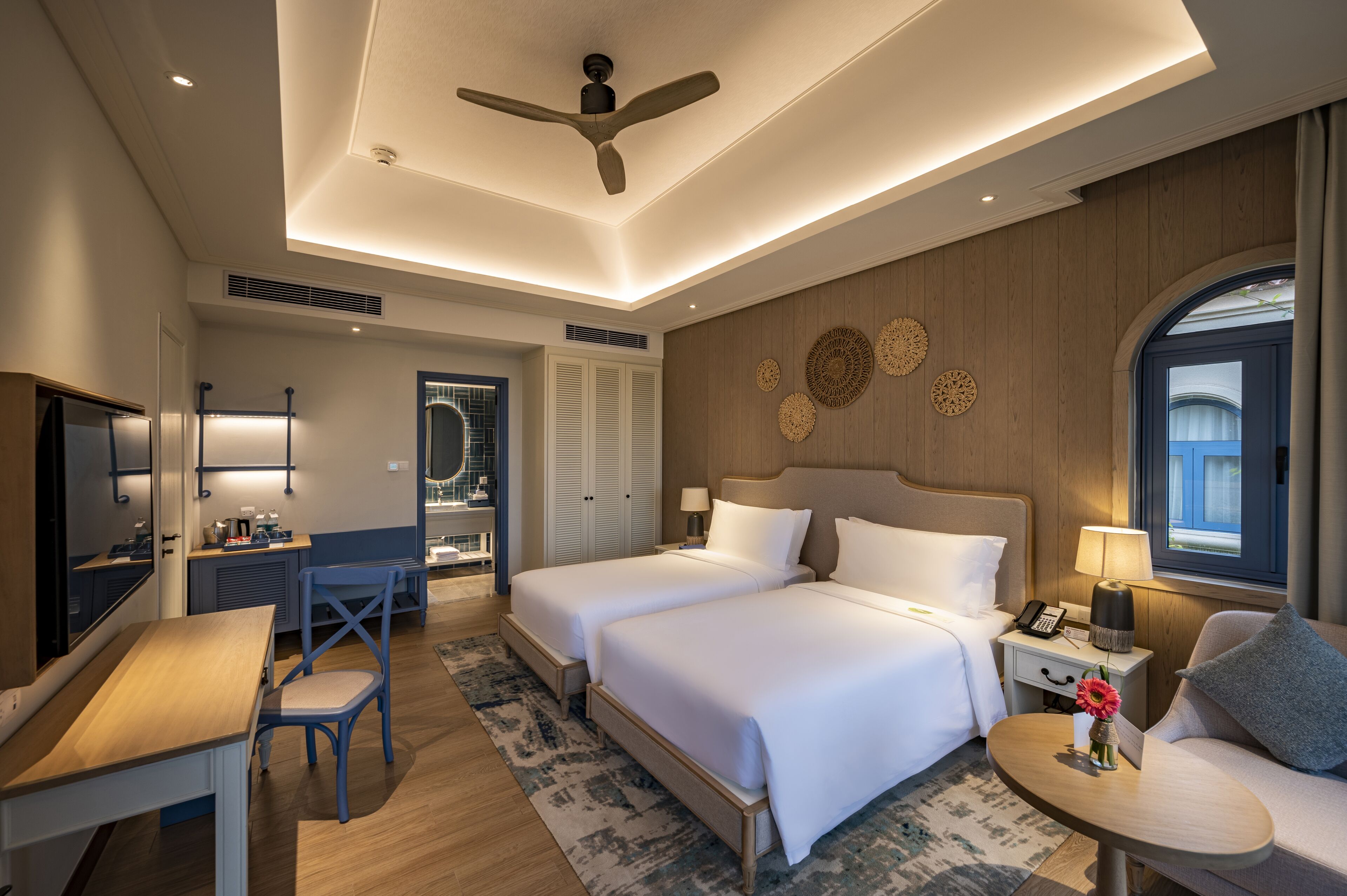 Une chambre d’hôtel avec deux lits, un bureau, une chaise, une petite table et un ventilateur de plafond.