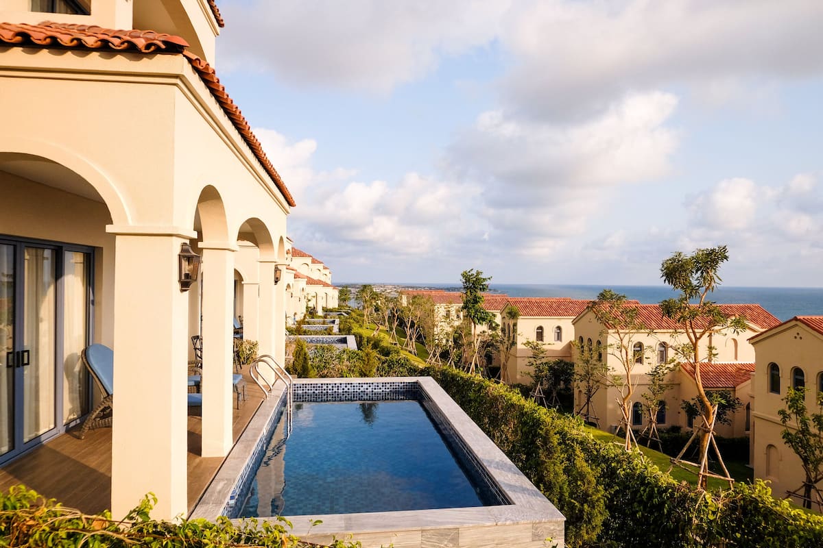 A swimming pool with a view of the sea, surrounded by buildings with arched windows and a tiled roof.