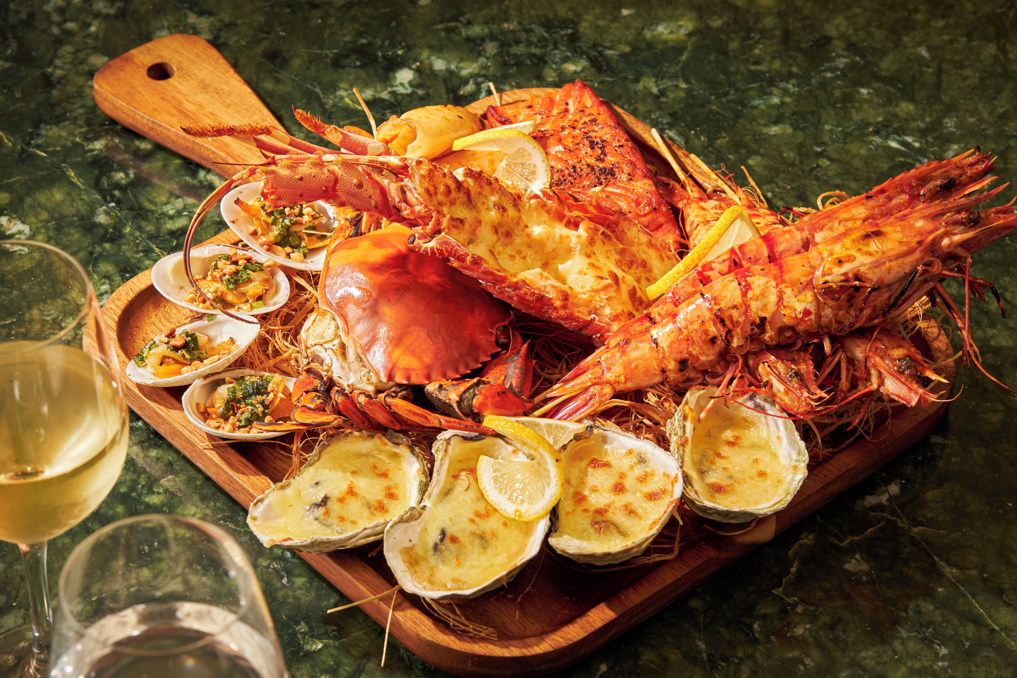 Una bandeja de madera con mariscos cocidos, incluyendo langostas y ostras, acompañada de rodajas de limón y una copa de vino blanco.