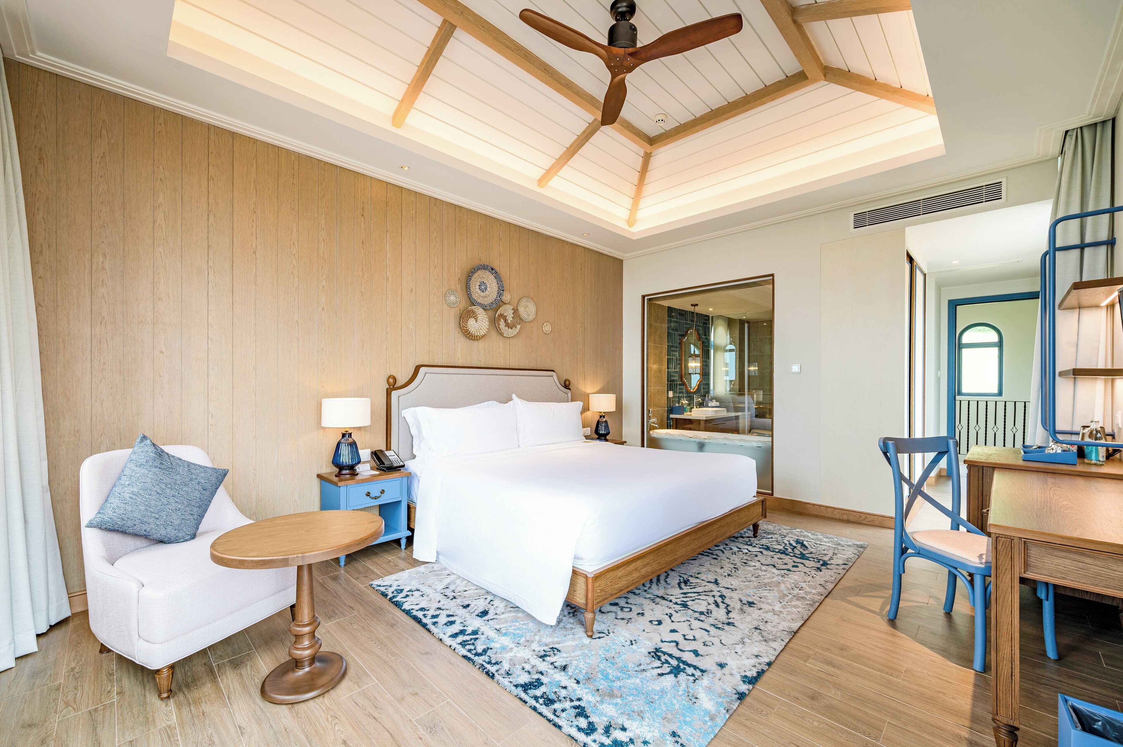 A hotel room with a large bed, a desk, a chair, a small table, and a ceiling fan.