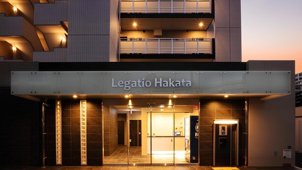 Esterno dell'hotel Legatio Hakata, dal design moderno, con un ingresso ben visibile, insegne illuminate e una vista chiara degli interni tramite le porte in vetro.