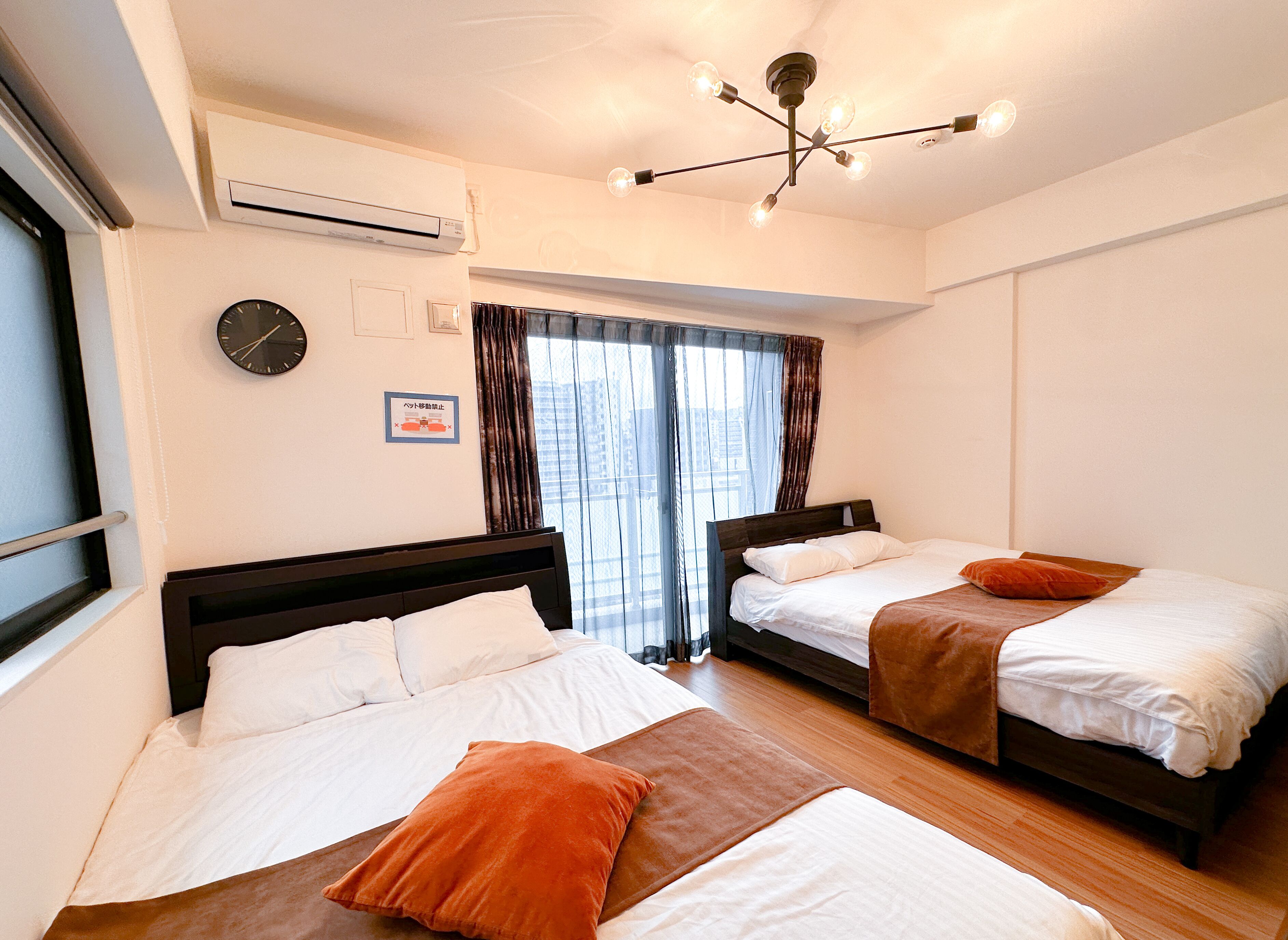 A hotel room with two beds, a ceiling light fixture, an air conditioning unit, a clock, and a window with curtains.