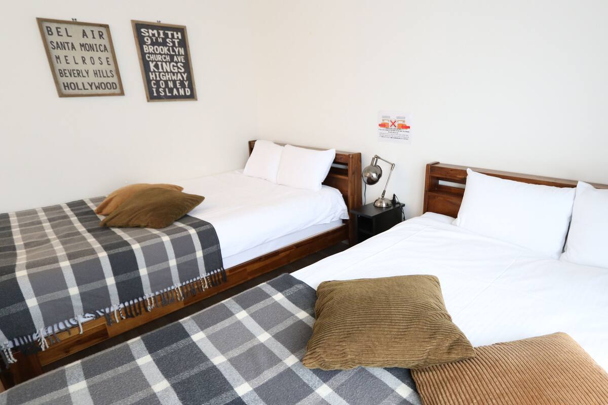 Habitación de hotel con dos camas, cada una con ropa de cama de estilo diferente. Hay dos letreros en la pared, una mesita con una lámpara y un cartel con una 'X' roja.