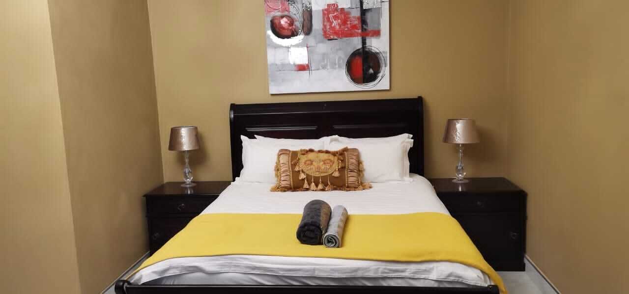 Une chambre d’hôtel avec un lit, deux tables de chevet, une lampe sur chaque table, une couvre-lit jaune, un oreiller blanc, un coussin décoratif et un tableau au mur.