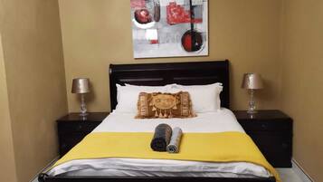 Une chambre d’hôtel avec un lit, deux tables de chevet, une lampe sur chaque table, une couvre-lit jaune, un oreiller blanc, un coussin décoratif et un tableau au mur.