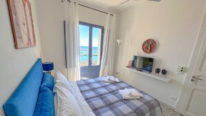 A hotel room with a bed, a TV, a window with curtains, a ceiling fan, and a view of the sea. - The fisherman's house (Sciacca)